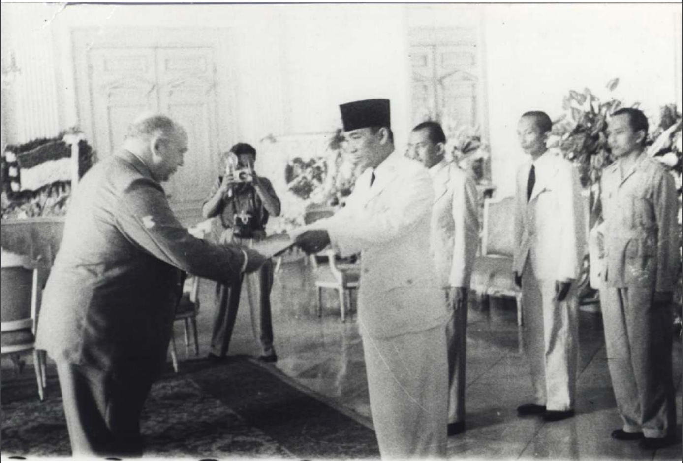 A historical black-and-white photo of a formal meeting, featuring two men exchanging documents, with others observing in a grand room.